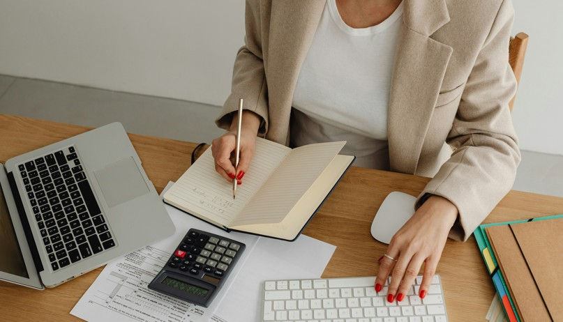 A person in a beige blazer writes in a notebook and uses a keyboard. A laptop, calculator, and files are on a wooden desk.