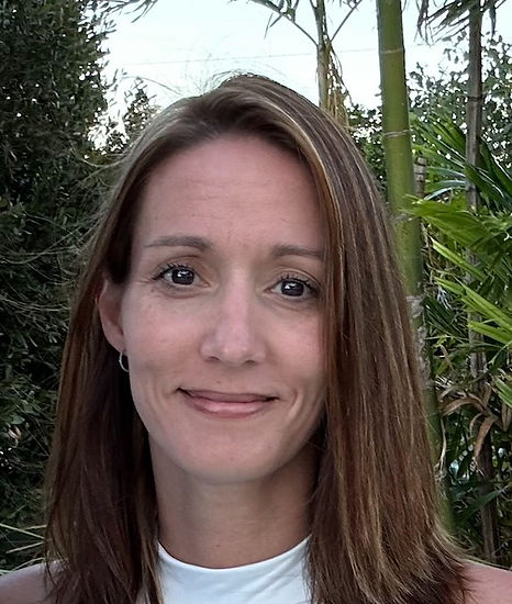 Smiling woman with long brown hair stands outdoors in a lush garden with palm trees and greenery.