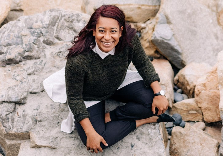 A smiling person with dark red hair sits on rocks wearing a dark sweater, white shirt, and black pants. Background features large stones.