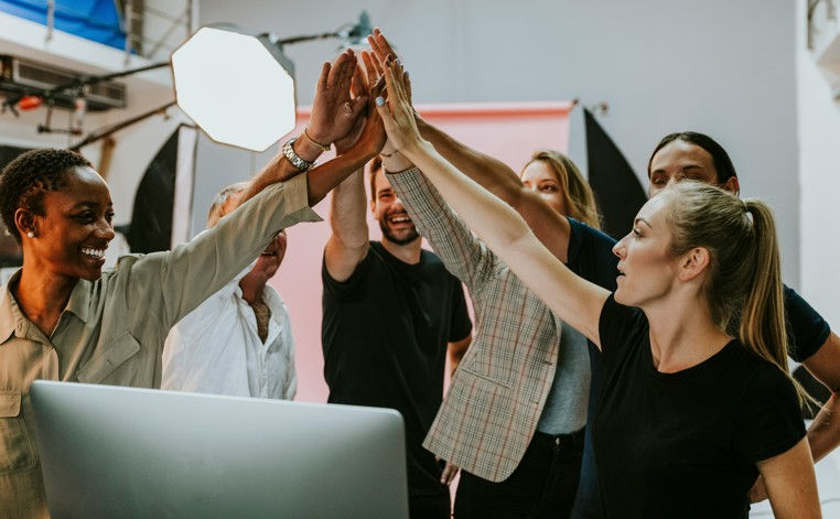 A group of diverse people smiling and giving a high-five in a studio setting with a computer and lighting equipment in the background.