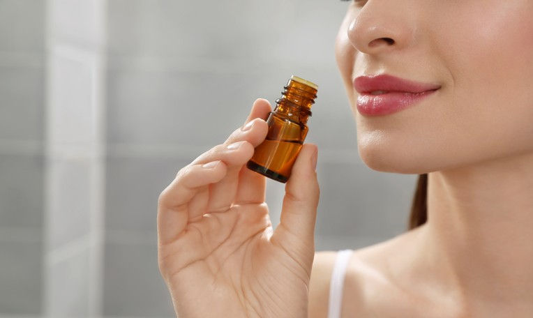 Woman holding a small amber bottle near her face, smiling slightly. Background is blurred and neutral, suggesting an indoor setting.