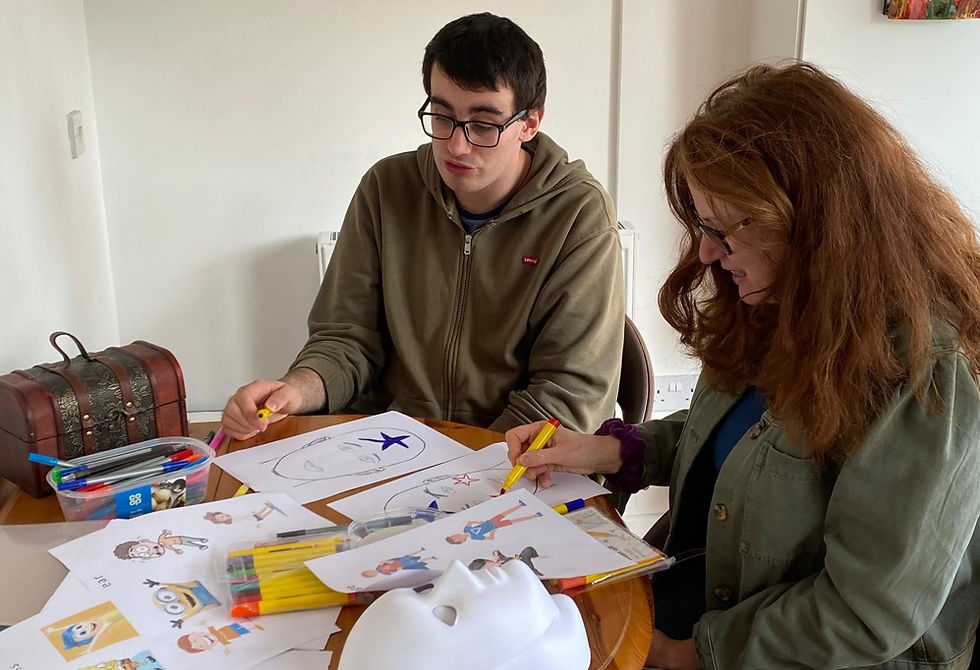 Two people sit at a table drawing with markers, surrounded by sketches and a white mask in a bright room.