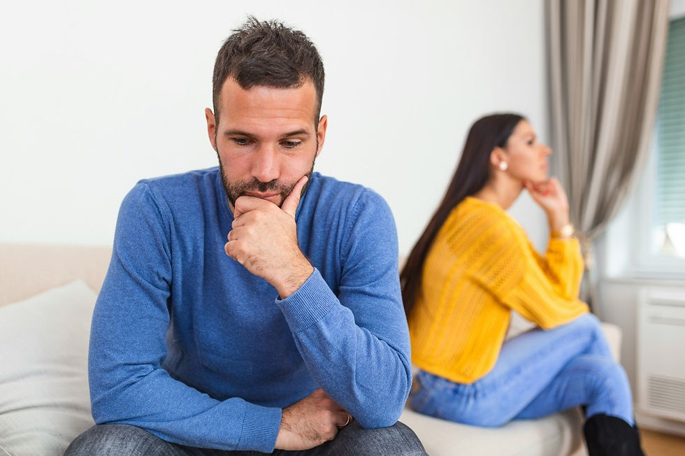 Thoughtful man in blue sweater sits foreground while woman in yellow sits apart in background, tense mood in a bright bedroom