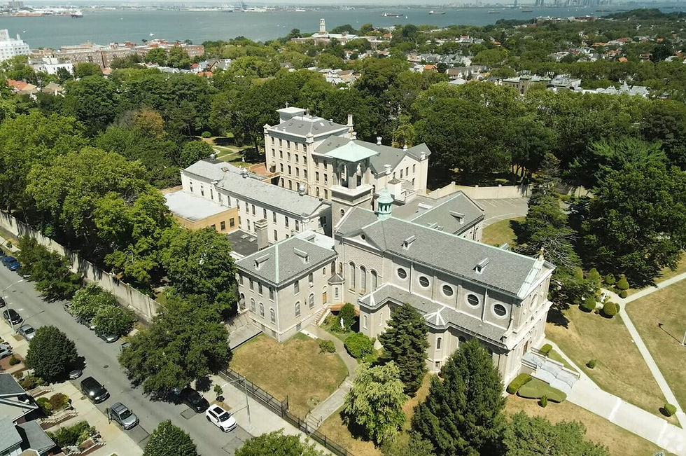 Aerial view of a historic stone complex with gray roofs in a green park near the waterfront and city skyline.
