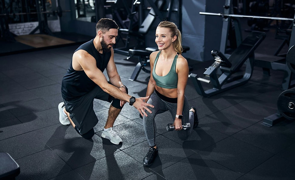 Personal trainer guides a smiling woman through a dumbbell lunge in a dark gym with exercise machines.