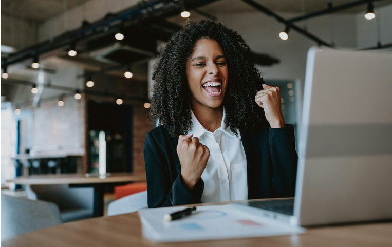 Woman with curly hair celebrates in front of a laptop in an office. She shows excitement with clenched fists. The room has industrial decor.