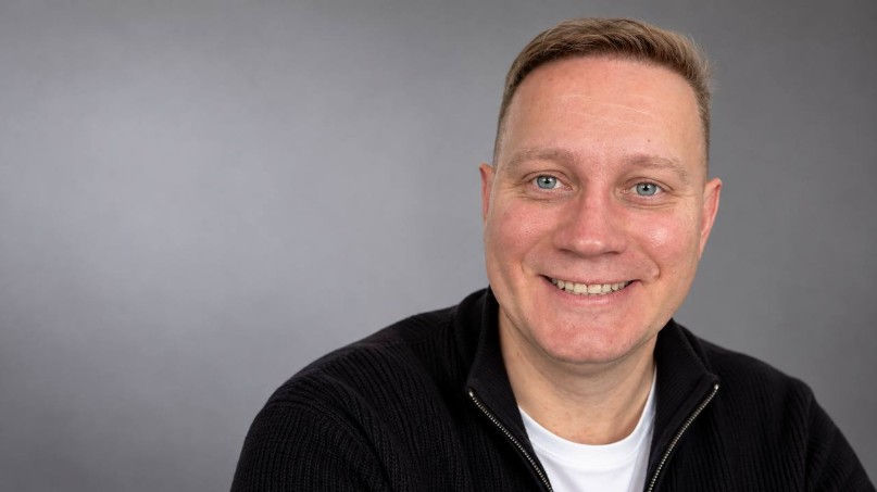 Smiling man in a black sweater against a plain gray background, posed in a studio-style portrait.