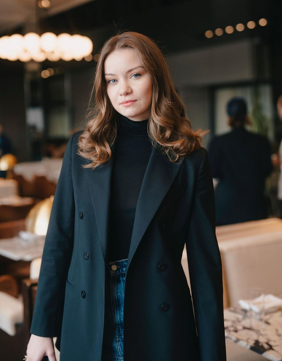 Woman in a black coat and jeans stands in a warmly lit restaurant, looking confidently at the camera. Background features tables and chairs.
