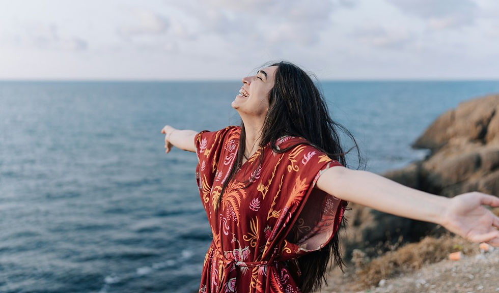 Woman in a red patterned dress stands on a rocky coast with arms outstretched, eyes closed, smiling at the sea.