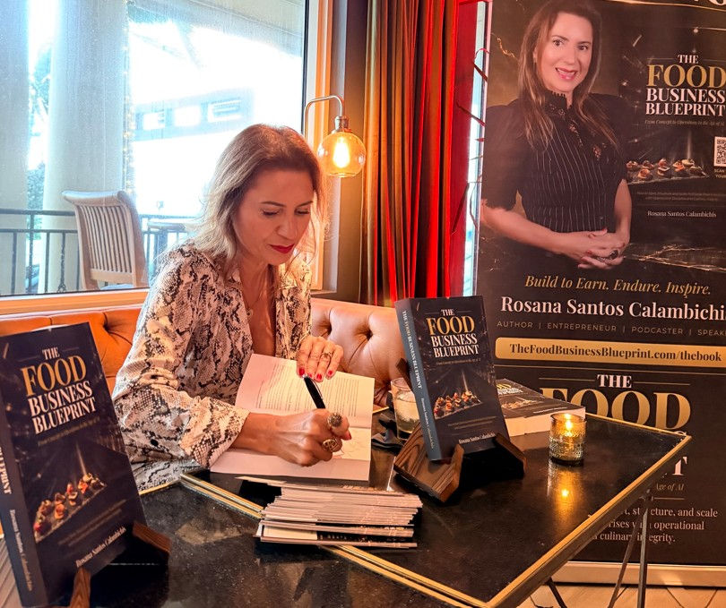 Author Rosana Santos Calambichis signs The Food Business Blueprint books at a cozy café table with posters and candles.