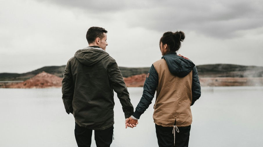Couple in jackets holding hands by a calm lake under a cloudy sky, standing together and facing the water