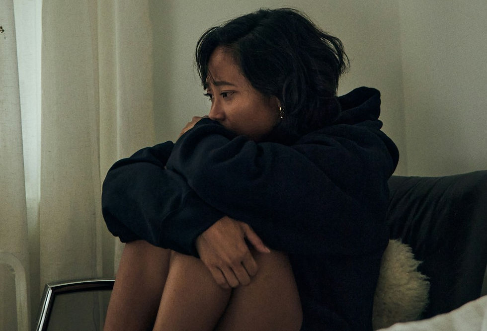Pensive woman in a black hoodie sits curled on a couch by a curtained window in a dim room, looking worried and withdrawn