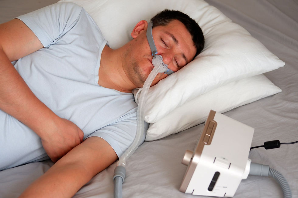 Man sleeping on a pillow with a CPAP mask, connected to a machine. He is wearing a light blue shirt, lying on a grey sheet.