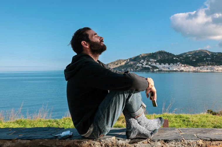 Man sitting on a stone ledge, eyes closed, holding a phone. Overlooks a calm sea and distant town, wearing a black hoodie and jeans.