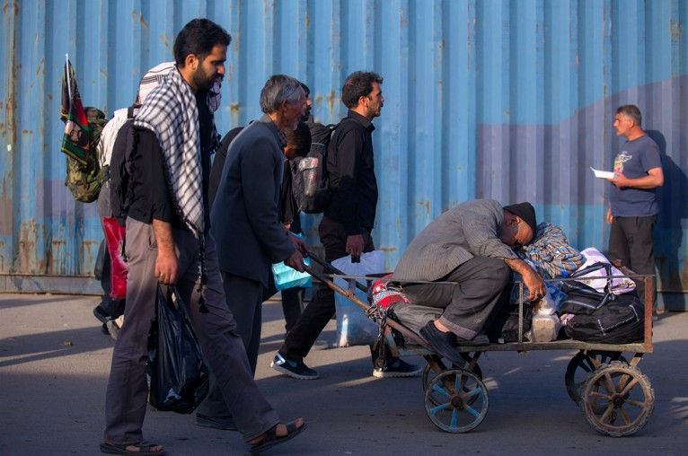 People walk beside a blue wall, pushing a cart filled with belongings. One person rests on the cart, appearing weary. Bright, sunny day.