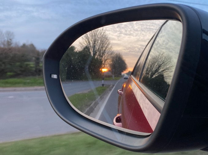 Car side mirror reflects a sunset over a quiet road lined with trees. The sky is textured and has soft colors, creating a calm mood.
