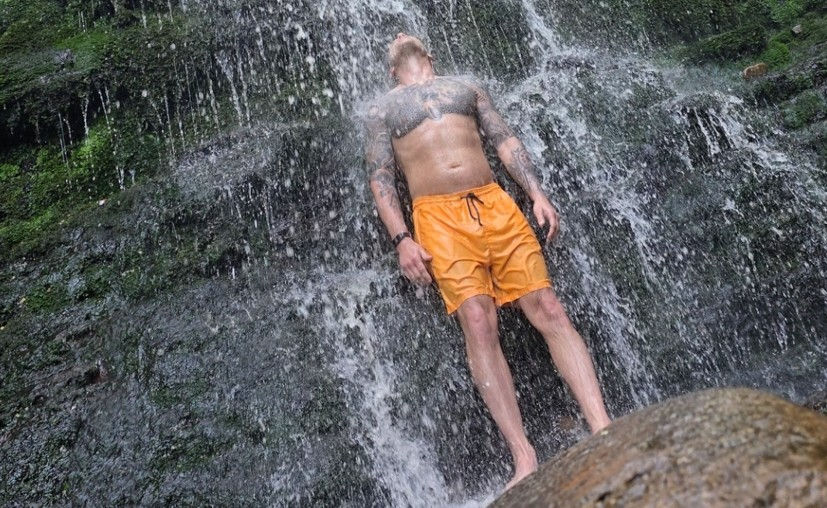 Tattooed man in orange shorts stands under a rushing waterfall against a rocky cliff, head tilted back, looking refreshed.