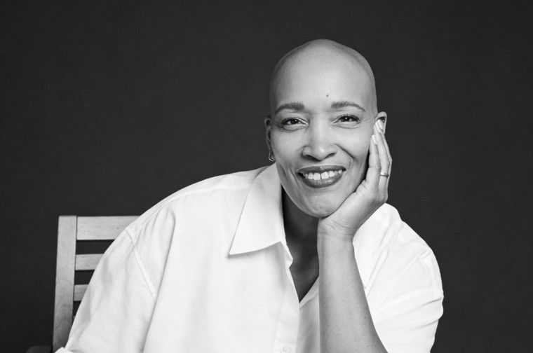 Smiling person in a white shirt rests their head on their hand, sitting on a wooden chair against a dark background. They appear relaxed and content.