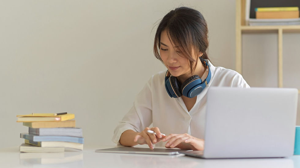 Woman working on a laptop at a desk with headphones around her neck, beside a stack of books in a calm home office.