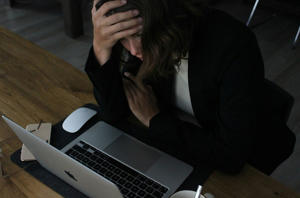 Stressed woman at a desk with a laptop and mouse, holding her forehead in a dim office, looking overwhelmed.