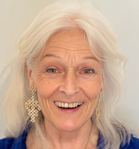 Elderly woman with long white hair, wearing ornate earrings and a blue top, smiling warmly against a plain light background.