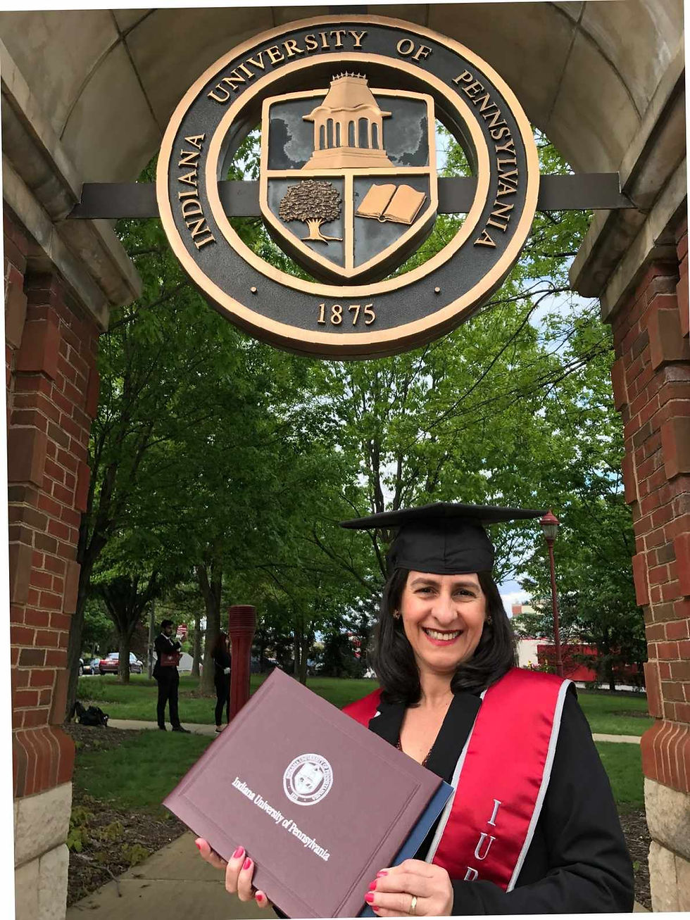 Graduate in cap and gown holds diploma, smiling under Indiana University of Pennsylvania arch. Red and black details, lush green background.