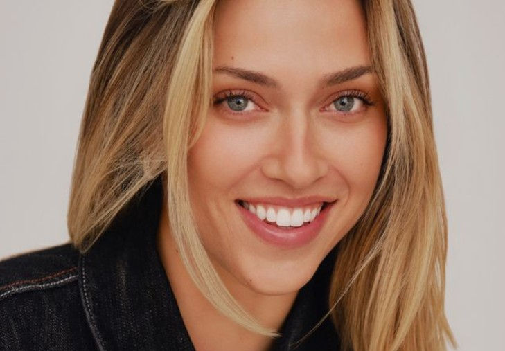 Smiling woman with blonde hair in a dark jacket, against a plain background. She has an expression of warmth and friendliness.