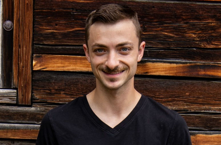 Man in black shirt smiles in front of a rustic wooden wall. Earthy tones create a warm, casual ambiance.
