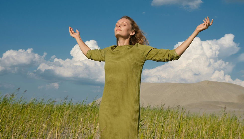 Woman in a green dress stands in a grassy field, arms raised and eyes closed, with dunes and clouds under a blue sky.