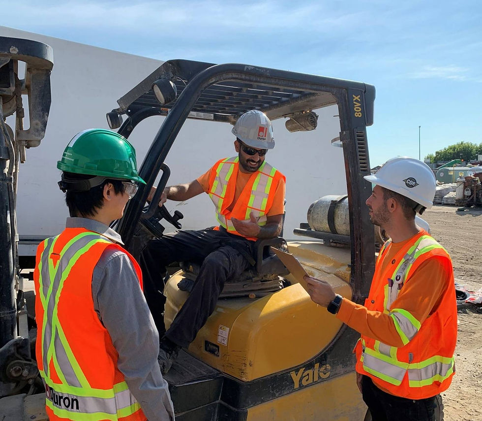 Three construction workers in safety gear discuss on a job site. One sits on a yellow forklift, others hold a clipboard. Sunny day.