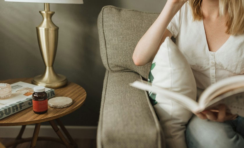 Woman in white top reads a book on a beige sofa beside a brass lamp and small table with a bottle, in a calm living room.