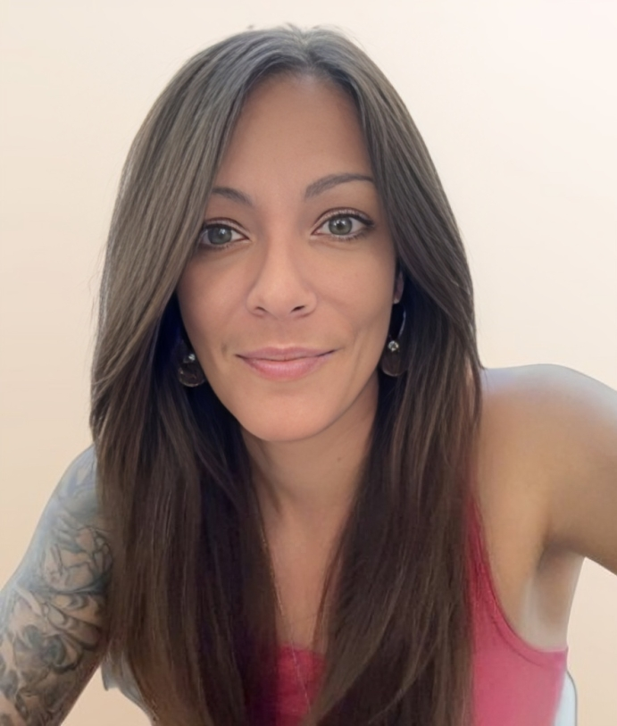 Smiling woman with long brown hair, wearing a pink tank top and earrings. Tattoo visible on arm. Neutral beige background.