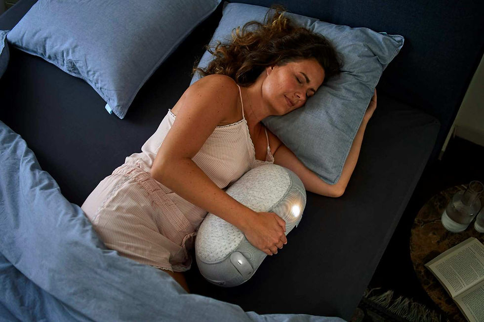 Woman in a beige top sleeping, hugging a soft, light-colored pillow on a bed with blue bedding. Relaxed mood, dimly lit room.
