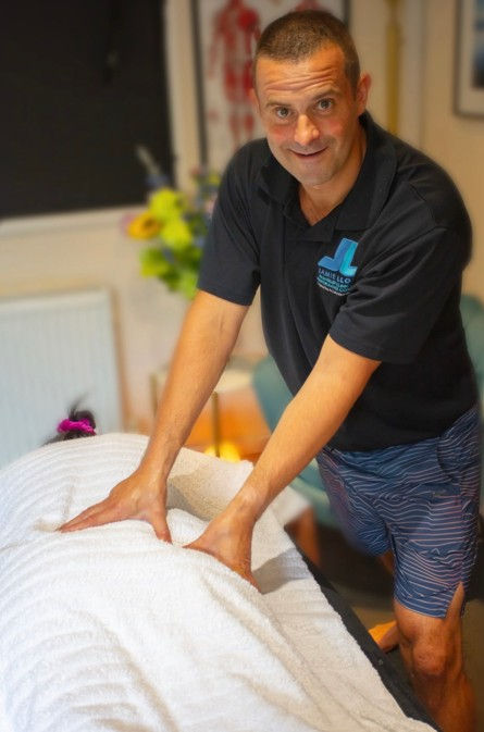 Man in a black polo gives a massage to a client under a white towel in a cozy room, smiling at the camera.