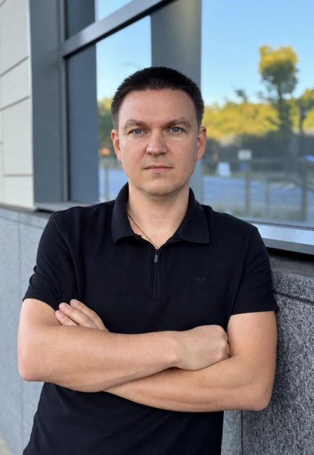 Man in a black polo stands with arms crossed by a window, looking serious, with trees and blue sky reflected behind him.