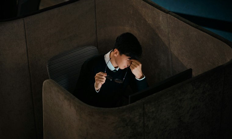 A person in a black sweater sits in a cubicle, holding glasses, looking stressed. Dim lighting creates a somber mood.