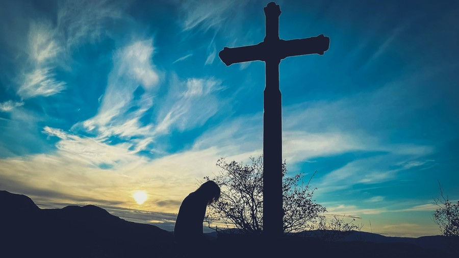 Silhouette of a bowed person beside a tall cross at sunset, with blue clouds and dark hills, conveying quiet reflection.