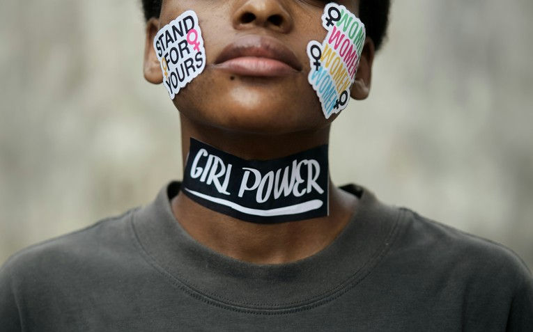 Close-up of a person with stickers reading "STAND FOR YOURS" and "GIRL POWER" on face and neck, wearing a dark shirt against a neutral background.