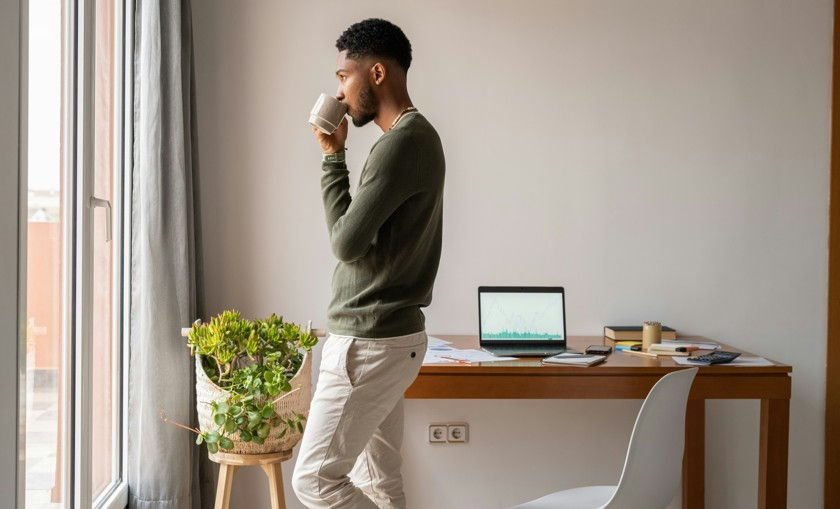 A man in a green sweater and light trousers stands by a large window, drinking from a mug and looking outside. In the background, a wooden desk holds a laptop displaying data charts, surrounded by papers and office supplies in a minimalist room.