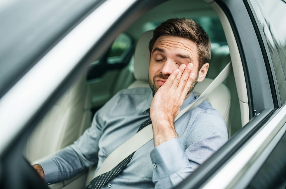 Tired man in a car rubs his eye while wearing a seatbelt, looking exhausted in the light-colored car interior.