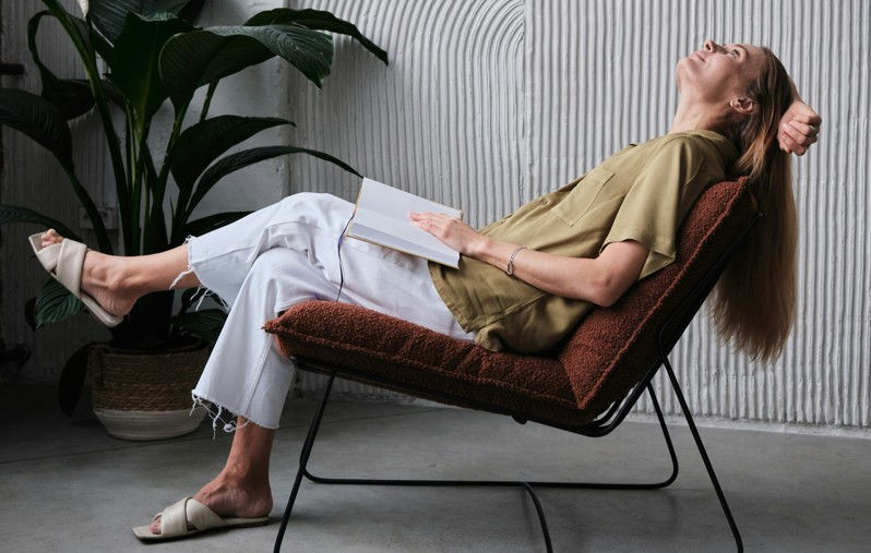 Woman in a relaxed pose on a chair, holding a closed book, against a textured wall and greenery. Warm tones create a calm mood.