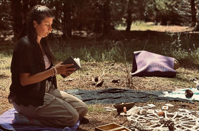 A woman kneels on a blanket in a forest, reading a book. Objects, including ropes and cards, are placed on the ground around her. Sunlight filters through trees.