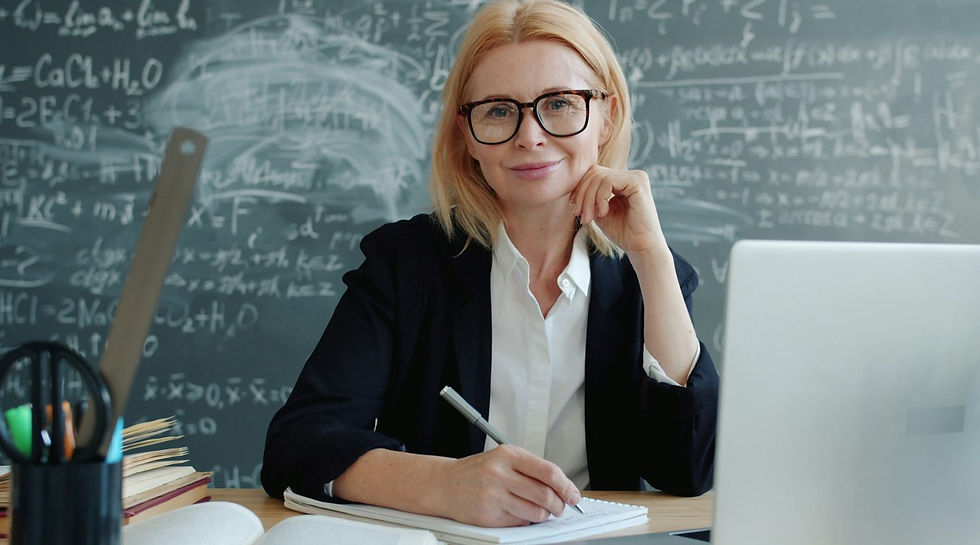Blonde teacher with glasses writes in a notebook at a desk, smiling in front of a chalkboard full of math formulas and a laptop.