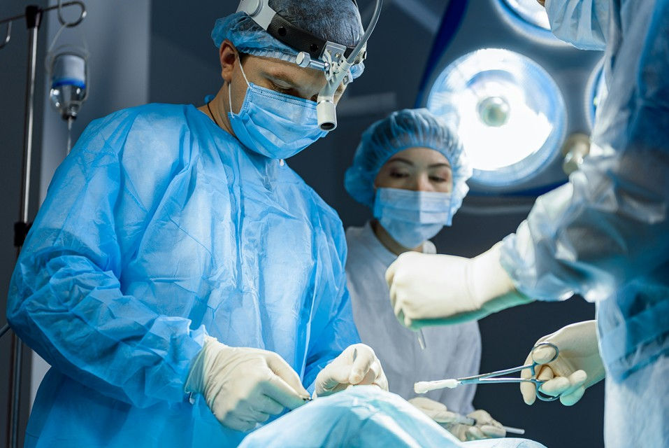 Surgeons in blue scrubs and masks perform an operation in a brightly lit operating room, focused and sterile.