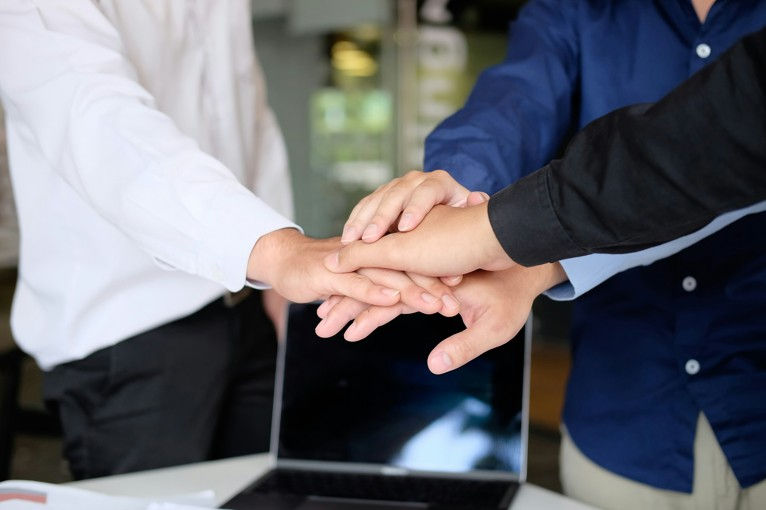 Three people stack hands over a laptop in an office, signaling teamwork and unity.