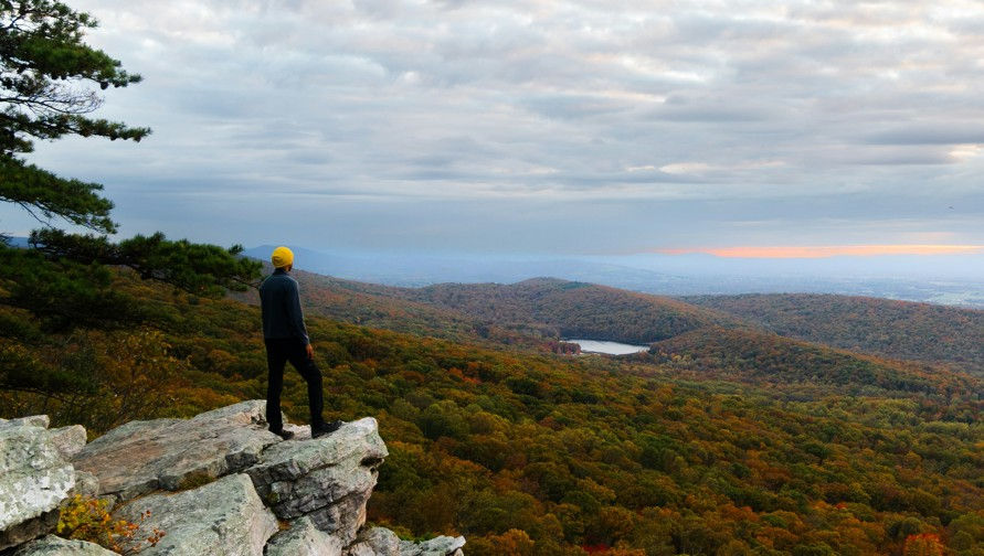 Person in a yellow beanie stands on a rocky cliff overlooking an autumn forest valley and lake under a cloudy sky.