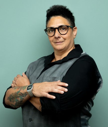 Person with glasses and tattoos, wearing a black and gray shirt, crosses arms confidently against a plain teal background.