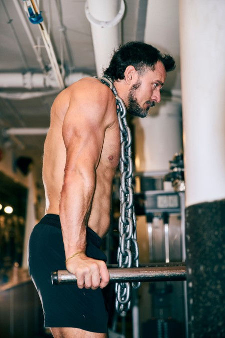 Shirtless man in a gym doing weighted dips, chain around his neck, focused and straining beside white columns.