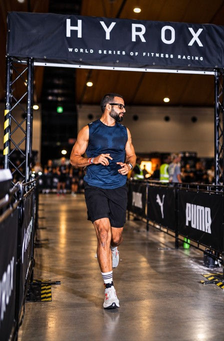 Athlete jogs under a HYROX World Series of Fitness Racing banner in an indoor arena, flanked by PUMA barriers.