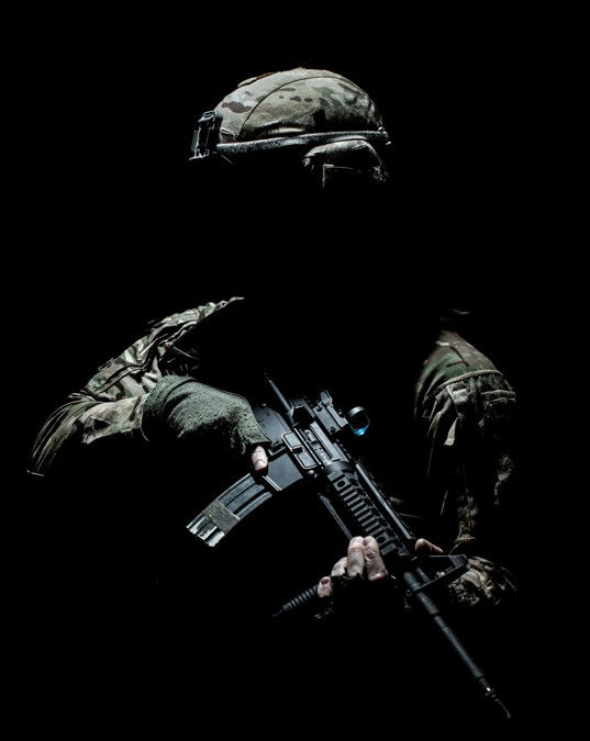 Camouflaged soldier in darkness holding a rifle, lit dramatically against a black background, tense and stealthy.